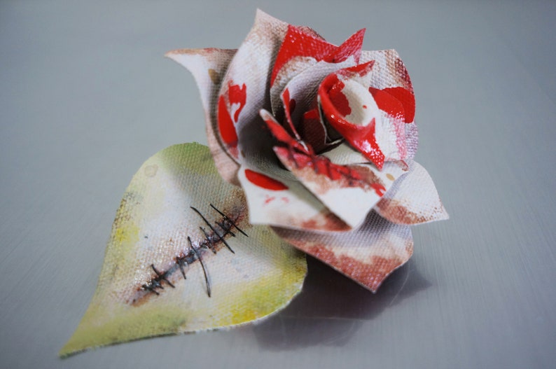 May include: A white fabric rose with red paint splatters and a stitched heart-shaped leaf. The rose is made of layered fabric and has a distressed look.