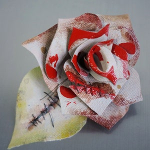 May include: A white fabric rose with red paint splatters and stitched details. The rose is made of fabric and has a distressed look.