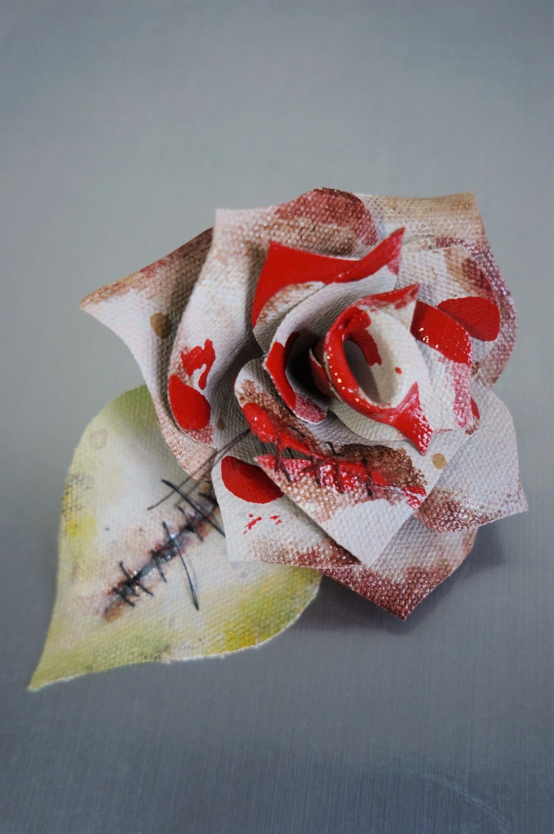 May include: A white fabric rose with red paint splatters and stitched details. The rose is made of fabric and has a distressed look.