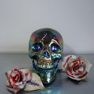 May include: A iridescent blue, green, and gold skull with a toothy grin sits on a gray surface with two white roses with red accents.