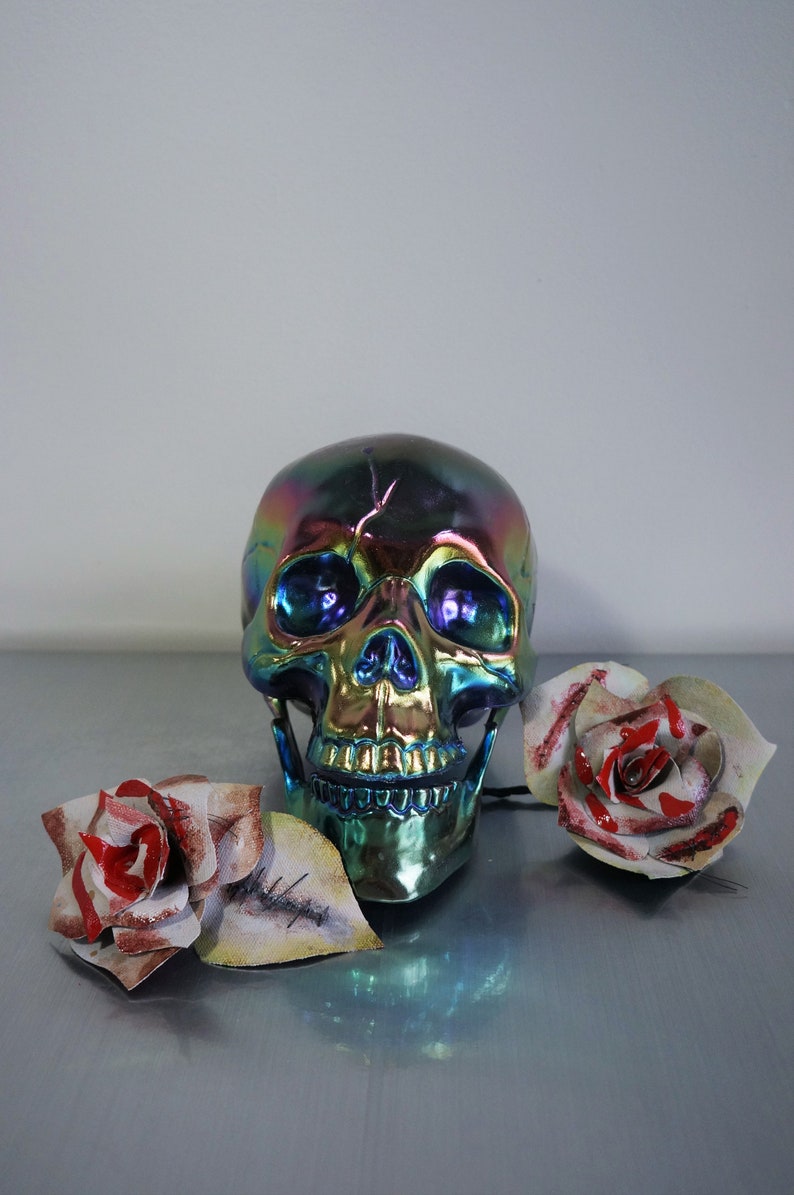 May include: A iridescent blue, green, and gold skull with a toothy grin sits on a gray surface with two white roses with red accents.