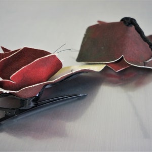 May include: A red fabric rose hair clip with a black elastic band. The rose is made of multiple layers of fabric and has a textured look.