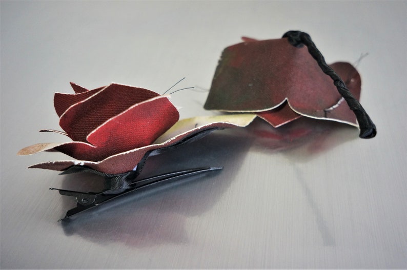May include: A red fabric rose hair clip with a black elastic band. The rose is made of multiple layers of fabric and has a textured look.