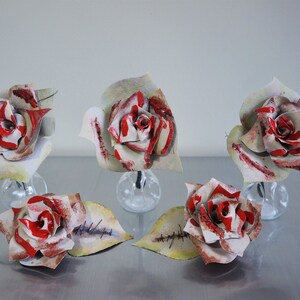 May include: Five paper roses in clear glass vases. The roses are white with red accents and have stitched details that look like wounds. The roses are arranged on a silver surface.