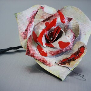 May include: A white fabric rose with red paint splatters and stitched details. The rose is attached to a black stem.