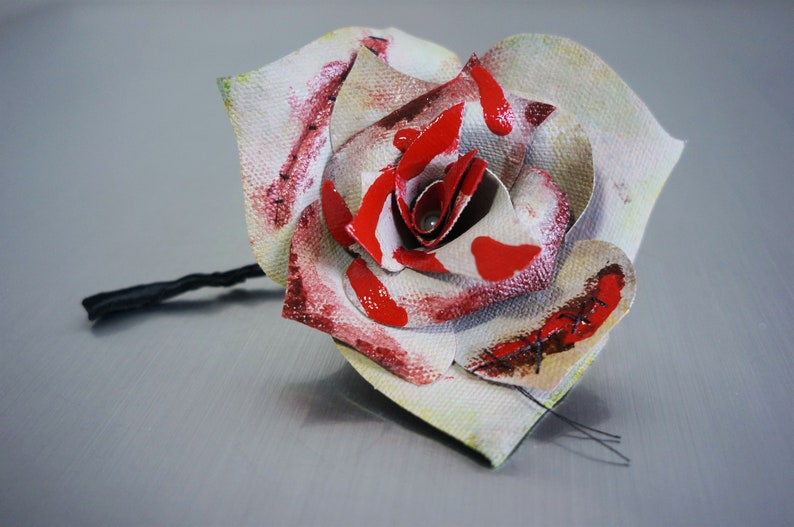 May include: A white fabric rose with red paint splatters and stitched details. The rose is attached to a black stem.