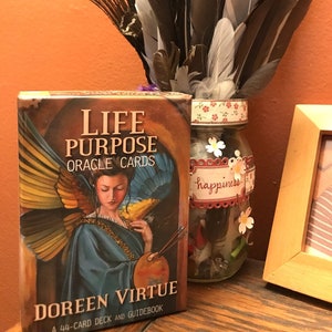 May include: A box of Life Purpose Oracle Cards by Doreen Virtue. The box features a painting of an angel with wings, holding a paintbrush and palette. The text "Life Purpose Oracle Cards" is printed in gold lettering on a brown background. The text "Doreen Virtue" is printed in white lettering on a blue background. The text "A 44-Card Deck and Guidebook" is printed in white lettering on a brown background.
