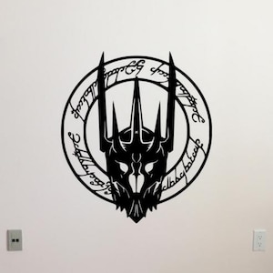 May include: Black metal wall art featuring a stylized, detailed depiction of a crown-like emblem within a circular frame. The design includes intricate details and script around the circle. The artwork is mounted on a white wall.