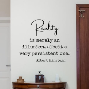 Puede incluir: Adhesivo de pared con la cita "La realidad es meramente una ilusión, aunque muy persistente" de Albert Einstein. El texto negro está sobre una pared blanca. Debajo de la cita hay una mesa de madera oscura con objetos decorativos.