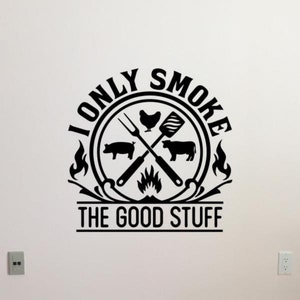 May include: Black and white graphic design with a circle containing a crossed fork, spatula, chicken, pig, and cow. The circle is surrounded by flames and the text "I Only Smoke The Good Stuff".