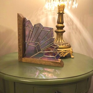 May include: A decorative stained glass piece with a geometric design in shades of purple and iridescent hues. The glass is set in a wooden frame. The piece is displayed on a green table, next to a gold lamp.