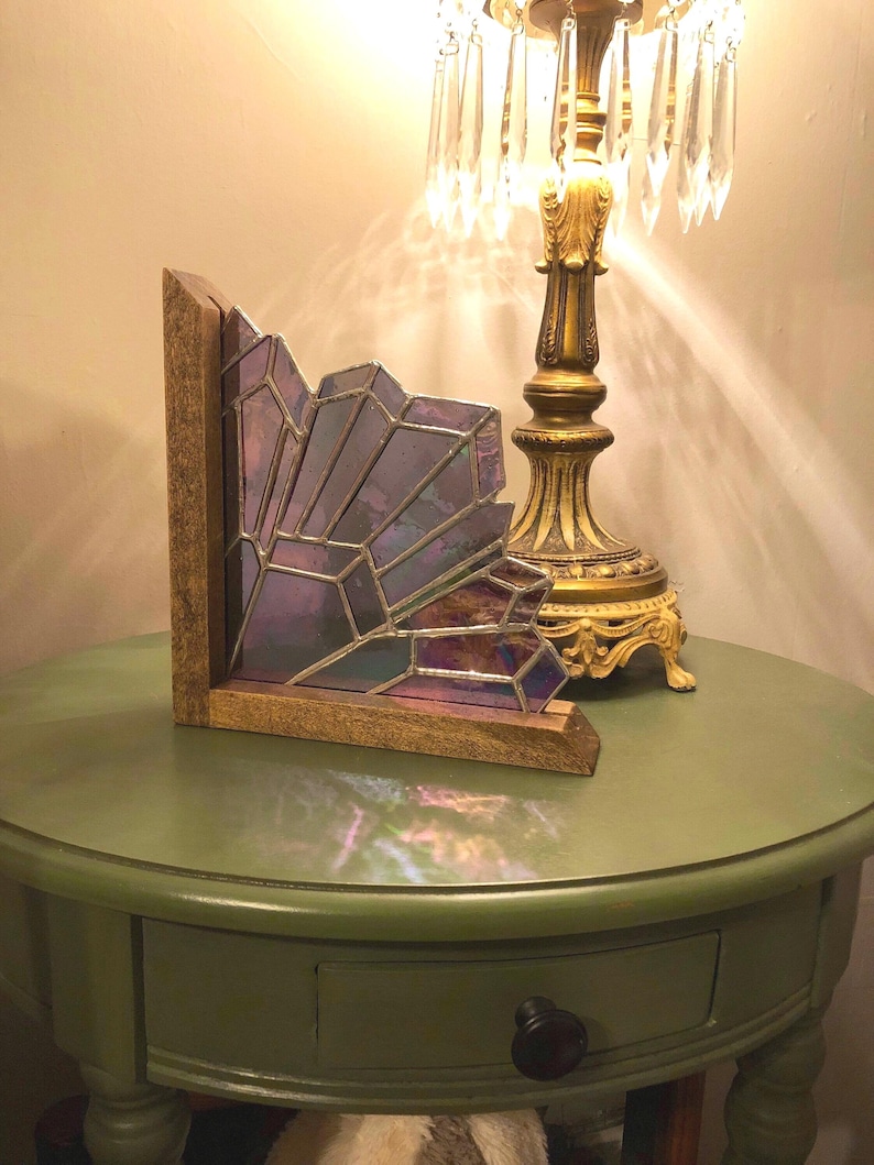 May include: A decorative stained glass piece with a geometric design in shades of purple and iridescent hues. The glass is set in a wooden frame. The piece is displayed on a green table, next to a gold lamp.