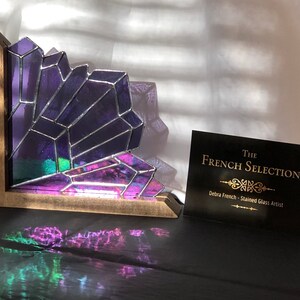 May include: A stained glass corner shelf featuring a geometric amethyst crystal design. The purple and iridescent glass is set in a wooden frame. A business card with the text "The French Selection" is also visible.
