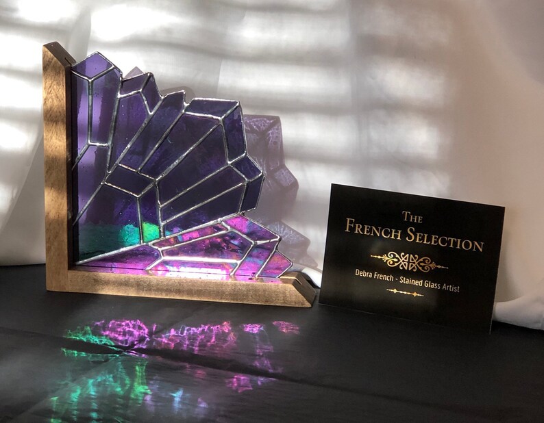 May include: A stained glass corner shelf featuring a geometric amethyst crystal design. The purple and iridescent glass is set in a wooden frame. A business card with the text "The French Selection" is also visible.