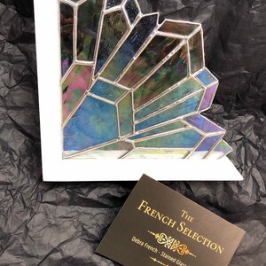 May include: A stained glass art piece with iridescent, geometric shapes in shades of blue, green, and purple, set within a white frame. The piece is a corner design. A business card reads "The French Selection - Debra French - Stained Glass Artist."