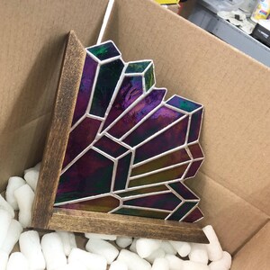 May include: A stained glass crystal sculpture with a wooden base. The glass features shades of purple, green, and gold, with white lines defining the geometric shapes. The sculpture is inside a cardboard box with packing peanuts.