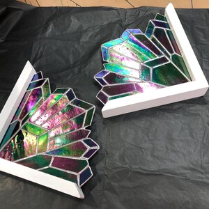 May include: Two decorative stained glass pieces in a geometric crystal design. The glass features iridescent colors of green, purple, and blue, set within a white wooden frame. The pieces are angled and appear to be corner accents.