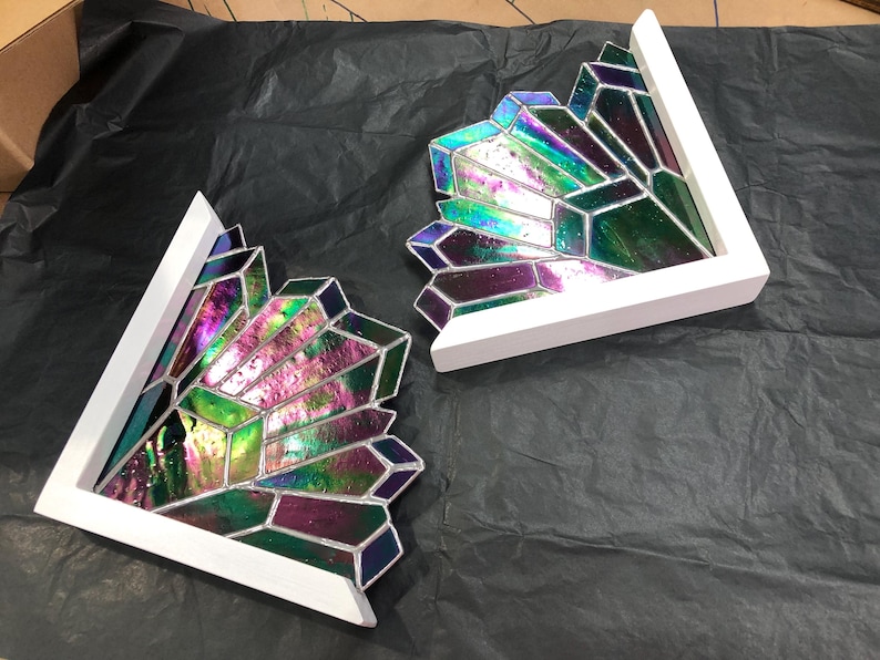 May include: Two decorative stained glass pieces in a geometric crystal design. The glass features iridescent colors of green, purple, and blue, set within a white wooden frame. The pieces are angled and appear to be corner accents.