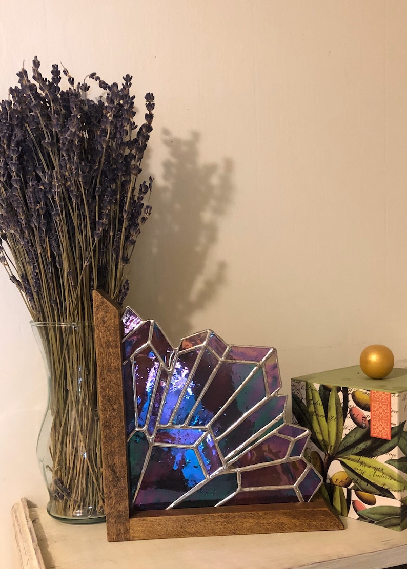 May include: A stained glass art piece with a geometric design in shades of purple, blue, and green, set in a wooden frame. A vase of dried lavender and a decorative box with a gold sphere are also in the image.