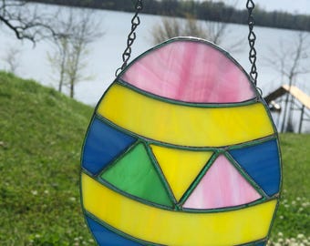 May include: A stained glass Easter egg ornament with a pink, yellow, blue, and green design. The egg is hanging from a silver chain.