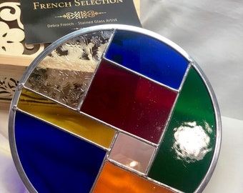 May include: A round stained glass piece with a silver frame. The glass is cut into geometric shapes and features a variety of colors, including blue, red, green, yellow, and orange.