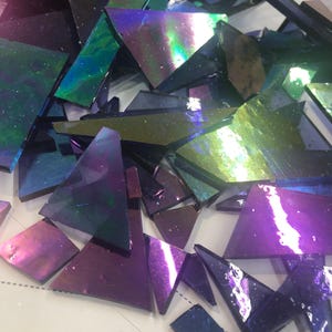 May include: A collection of iridescent glass pieces in various shapes and sizes. The glass shards display a range of colors, including purple, blue, green, and hints of gold. The pieces are scattered, suggesting they are for a mosaic or craft project.