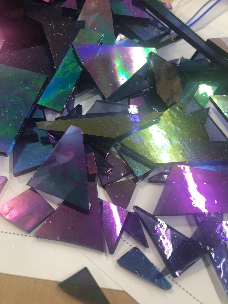 May include: A collection of iridescent glass pieces in various shapes and sizes. The glass shards display a range of colors, including purple, blue, green, and hints of gold. The pieces are scattered, suggesting they are for a mosaic or craft project.