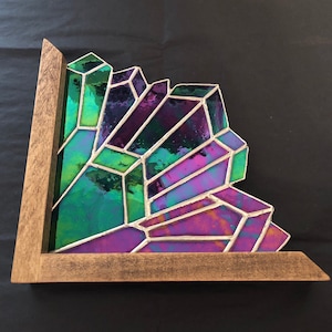 May include: A stained glass art piece featuring a cluster of geometric shapes in shades of green, purple, and blue. The glass is framed in a light brown wooden frame, creating a unique decorative accent.