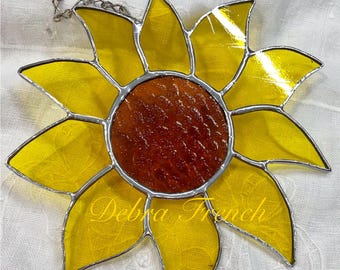 May include: A stained glass sunflower with yellow petals and a brown center. The petals are outlined in silver and the piece has a silver chain for hanging. The name "Debra French" is written on the bottom petal.