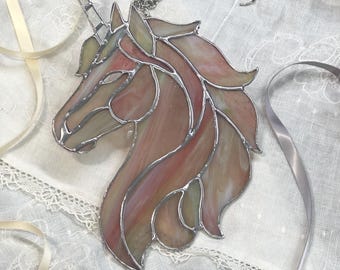 May include: A stained glass unicorn head suncatcher featuring iridescent pink, peach, and cream glass segments joined by silver metal solder lines. The piece includes a silver metal chain and hook attached to the top for hanging display purposes.