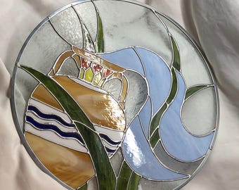 May include: A round stained glass piece featuring a stylized design. The artwork includes a yellow and white striped orb, blue and green flowing shapes, and a light blue element. The piece is framed with a silver metal border.