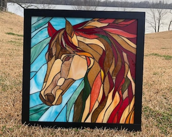 May include: Stained glass artwork of a horse's head in a black frame. The horse features a brown and tan face, with a flowing mane of red, orange, and brown. The background is a mix of blue and green.