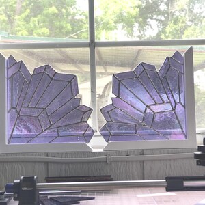 May include: Two stained glass panels in a window, each with a jagged, crystal-like design. The glass is a mix of purple and iridescent colors, set in a white frame. The window is in front of a blurred outdoor scene.