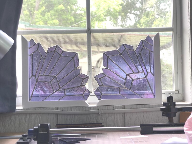 May include: Two stained glass panels in a window, each with a jagged, crystal-like design. The glass is a mix of purple and iridescent colors, set in a white frame. The window is in front of a blurred outdoor scene.
