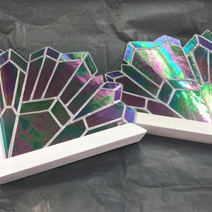 May include: Two white-framed, L-shaped decorative pieces. Each features a geometric design of iridescent glass in shades of purple, green, and blue. The glass is segmented with silver-colored lines, creating a crystalline appearance. The pieces are set against a dark background.