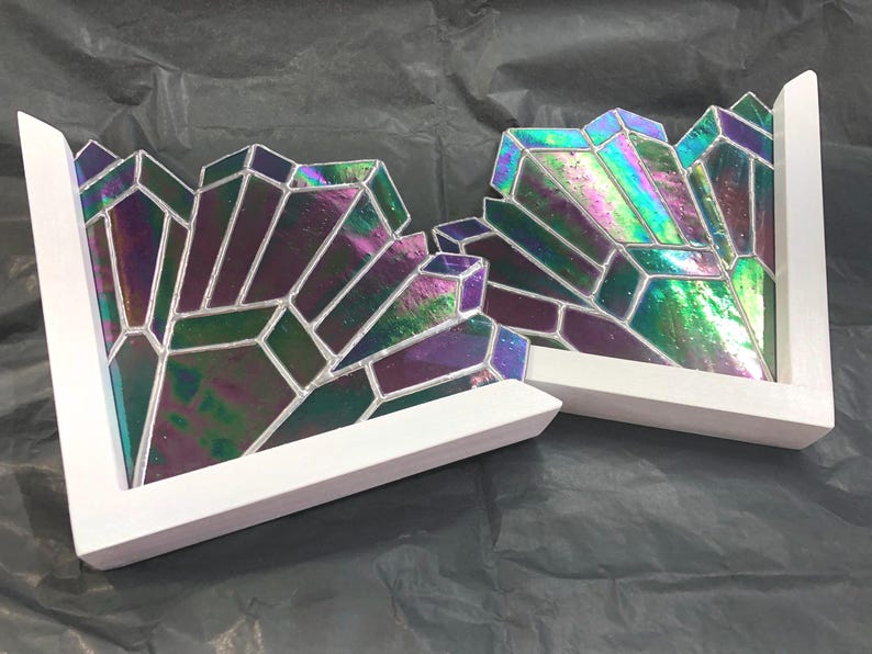 May include: Two white-framed, L-shaped decorative pieces. Each features a geometric design of iridescent glass in shades of purple, green, and blue. The glass is segmented with silver-colored lines, creating a crystalline appearance. The pieces are set against a dark background.