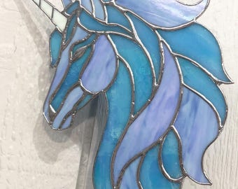 May include: A stained glass unicorn head suncatcher featuring shades of light blue and lavender glass pieces held together by silver metal solder lines. The unicorn has a long, pointed white horn extending from its forehead and a flowing mane.