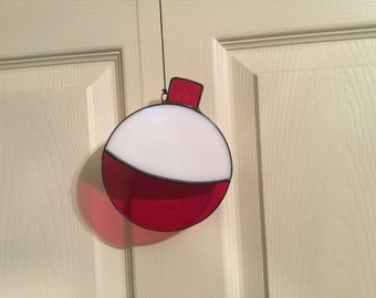May include: A stained glass fishing bobber ornament. The ornament is round with a white top and a red bottom section. A small red rectangular piece is at the top. The bobber is hanging from a black string.