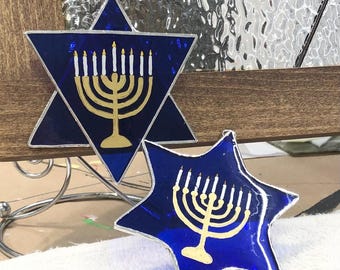May include: Two blue Star of David ornaments with a gold menorah design. The menorah has nine candle holders with white candles. One ornament is a six-pointed star, and the other is a five-pointed star.