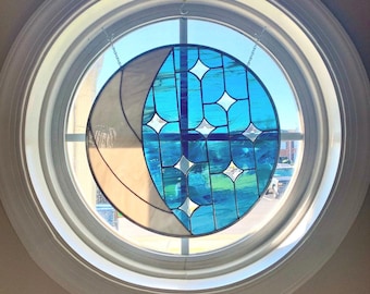 May include: A round stained glass window with a celestial design. The window features a crescent moon shape in beige and a blue oval with white diamond-shaped stars. The window is set in a white circular frame.