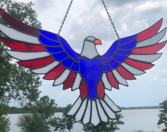 May include: A stained glass suncatcher shaped like an eagle with wings spread wide. The eagle features red, white, and blue glass panels outlined in dark metal solder, with a grey head and an orange beak, suspended by a thin metal chain.
