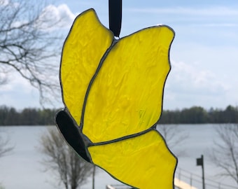 May include: A yellow stained glass butterfly hanging from a black ribbon. The butterfly is in a side view with the wings spread out. The background is a blurry image of a lake and trees.