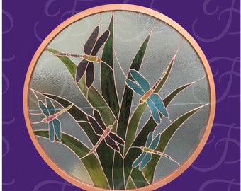 May include: A circular stained glass panel featuring four dragonflies in shades of blue and dark purple, surrounded by long green leaves. The design is encased in a smooth copper-colored metal frame, creating a decorative piece of art.