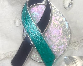 May include: Teal and black ribbon made of stained glass with a iridescent background. The ribbon is hanging from a silver chain.