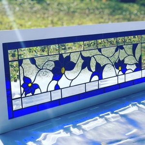 May include: A rectangular stained glass window with a white frame. The design features blue flowers with yellow centers and clear glass, outlined with black lines. The window is approximately 30 inches long.