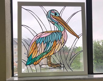 May include: A stained glass panel in a white frame featuring a pelican with multicolored wings in shades of blue, pink, and orange. The bird is depicted standing among tall, clear textured glass reeds, with black lead lines defining the shapes.