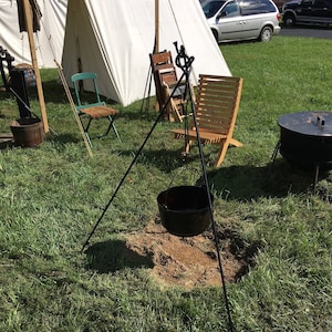May include: A black cast iron pot hanging over a fire pit with a tripod stand. The pot is hanging over a dirt fire pit in a grassy area. There are two wooden chairs and a white tent in the background.