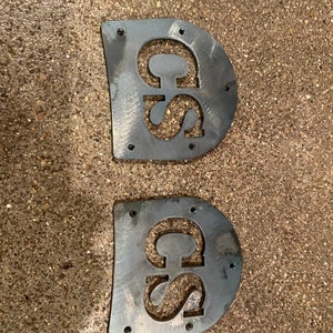 May include: Two metal cutouts of the letters "CS" in a rounded rectangular shape. The cutouts are made of a dark metal and have a rough, unfinished texture. There are several small holes around the edges of each cutout.