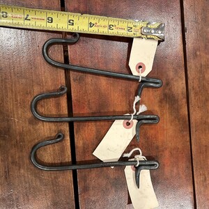 May include: Three hand-forged, dark gray metal hooks with a curved design. Each hook has a tag attached with string. The hooks are approximately 4 inches long, and are displayed on a wooden surface.