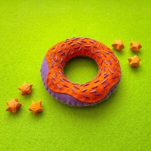 Peut inclure: Un beignet en feutre violet et orange avec des décorations en forme d'étoile orange sur un fond vert.
